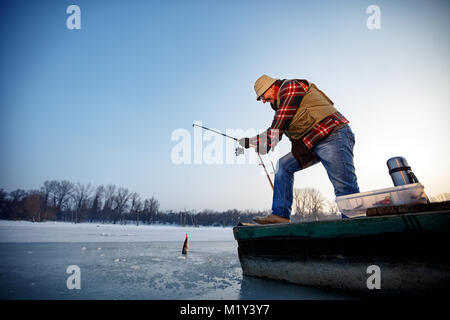 Lächelnd alter Fischer fangen Fische auf dem gefrorenen Fluss im Winter Stockfoto