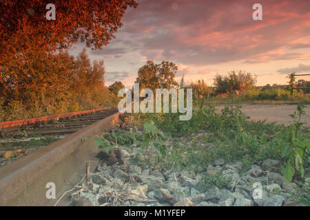 Im Vordergrund sind Steine, Bäume und Büsche. Im Hintergrund sind rot beleuchtet Wolken bei Sonnenuntergang. Eisenbahnschienen laufen hinter dem Horizont. Stockfoto