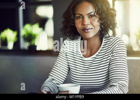 Junge afrikanische Frau selbstbewusst lächelnd, während allein sitzen an einem Tisch in einem Café genießen Sie eine frische Tasse Kaffee Stockfoto