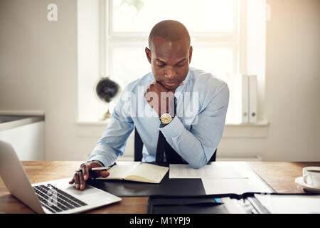 Junge afrikanische executive Lesen von Dokumenten und arbeitet an einem Laptop, während an seinem Schreibtisch in einem Büro sitzen Stockfoto