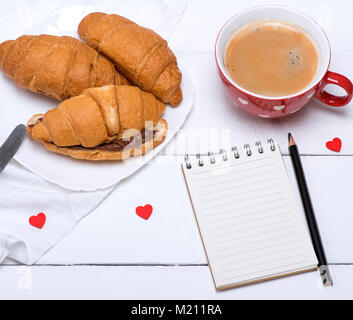 Rot Keramik Becher mit heißem Kaffee und frischen Croissants mit Schokolade, in der Mitte eine leere Notepad in Linie und schwarzem Holz Bleistift, Ansicht von oben Stockfoto