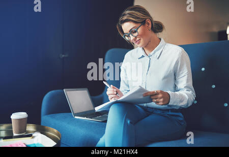 Lächelnden jungen Geschäftsfrau lesen durch Dokumente und ein Laptop verwenden, während auf einem Sofa zu Hause sitzen Stockfoto