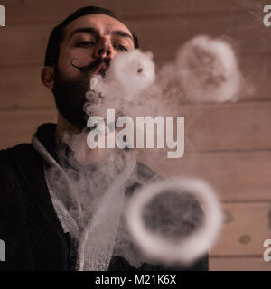 Mann mit Bart und Mustages Vaping eine elektronische Zigarette. Vaper Hipster Rauch Vaporizer und Exhals Rauch Ringe. Stockfoto