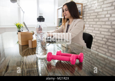 Wasserflasche und Rosa Hanteln auf Büro Schreibtisch mit einer Geschäftsfrau im Hintergrund Stockfoto