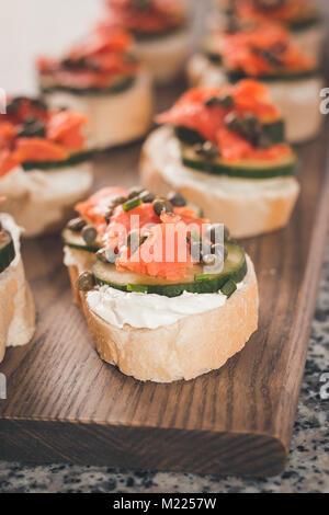 Mehrere finger Sandwiches mit geräuchertem Lachs und Frischkäse decken ein Schneidebrett. Kapern und in Scheiben geschnittene Gurken sind auf die Sandwiches enthalten. Stockfoto