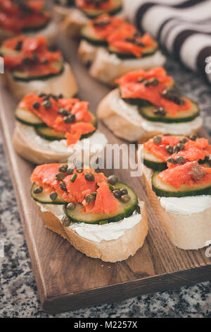 Mehrere finger Sandwiches mit geräuchertem Lachs und Frischkäse decken ein Schneidebrett. Kapern und in Scheiben geschnittene Gurken sind auf die Sandwiches enthalten. Stockfoto