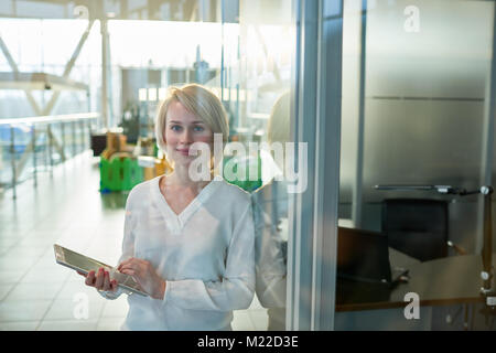 Lächelnde blonde white collar worker posieren für Fotografie halten digitale Tablett in den Händen und stehen im geräumigen Büro Lobby, Aussicht trou Stockfoto