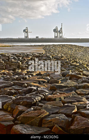 Schwere Steine, die Bestandteil des wellenbrechers am Strand im Aberavon mit drei großen Kräne in Port Talbot Stahlwerk Docks im Hintergrund. Stockfoto