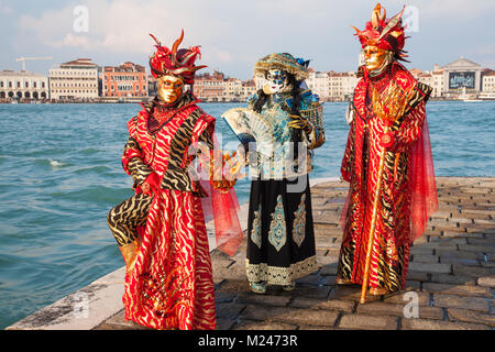 Venedig, Venetien, Italien 4. Februar 2018. Drei Menschen tragen bunte historische Kostüme und Masken im San Giorgio Maggiore mit der Lagune hinter ihnen an der Karneval von Venedig das Posieren. Stockfoto