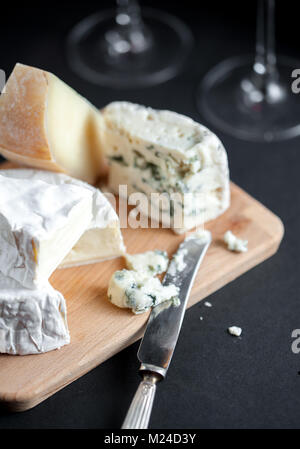 Auswahl an Käse auf einer Holzplatte mit Vintage silver Messer und zwei Weingläser. Vertikale. Makro Bild. Schwarzen Hintergrund. Vertikale. Stockfoto