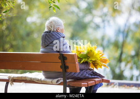 Ältere Frau auf Bank im Park sitzen und halten Haufen des Herbstes lässt Stockfoto