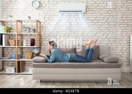 Liegende Frau mit Laptop auf dem Sofa genießen die Kühlung der Klimaanlage auf Home Stockfoto