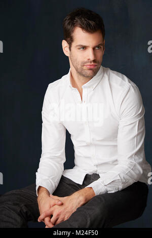 Studio Portrait von gutaussehenden jungen Mann tief in Gedanken tragen Shirt bei dunklem Hintergrund sitzen. Stockfoto