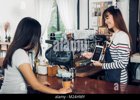 Asiatische Frau barista Schürze tragen Jean Vorschlag/Menü, um Kunden an Theke mit Smile Emotion, Cafe Restaurant Service Konzept, Inhaber kleine b Stockfoto