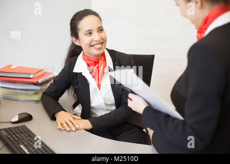 Portrait von Mixed-race Geschäftsfrau mit schwarzen und roten Uniform sitzen am Schreibtisch im Büro und freundlich lächelnd, während Kollege suchen Stockfoto