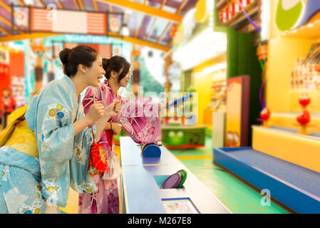 Fröhliche junge Frau traditionellen Kimono Kleidung zusammen tragen und spielen Daruma Spiel in Markt mit selektiven Fokus Foto. Stockfoto