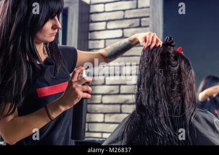 Schönes Haar stylist mit tätowierten Händen Anwendung Haarspray auf die Haare der Jungen brünette Frau, während sie sitzt in einem Sessel im Beauty Salon Stockfoto