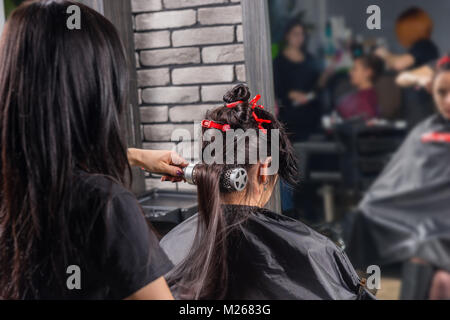 Weibliche Friseur putzen und Trocknen mit der Hand gehalten Fön auf die Haare der Jungen brünette Frau, während sie im Sessel sitzt im Schönheitssalon Stockfoto