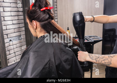 Weibliche Friseur mit tätowierten Hände putzen und Trocknen mit der Hand gehalten Fön auf die Haare der Jungen brünette Frau, während sie im Sessel sitzt. Stockfoto