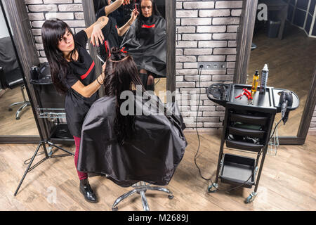 Blick von oben auf die weiblichen Friseur schneiden und Modellierung braunes Haar von Schere und Kamm von Junge brünette Frau, während sie im Sessel sitzt in ist Stockfoto