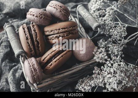 Frisch gebackene Makronen in Weidenkorb mit Griffen mit kleinen weißen Blüten auf Holz- Hintergrund. Stockfoto