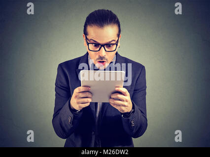 Formale Mann, verblüfft Sie Nachrichten auf Tablet und auf grauem Hintergrund posiert. Stockfoto