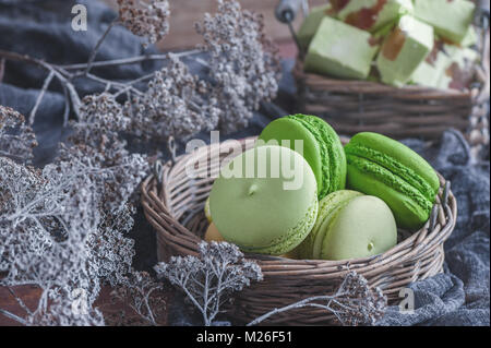 Frisch gebackene farbige Makronen in Weidenkorb mit Griffen mit kleinen weißen Blüten auf Holz- Hintergrund. Stockfoto