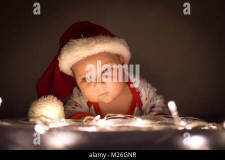 Cute Baby mit Weihnachtsmütze hob den Kopf über die Lichter unter dem Weihnachtsbaum