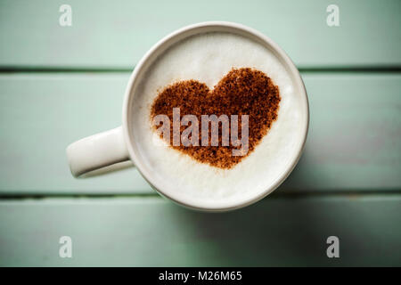 High-Angle Shot eine weiße Keramik Tasse Cappuccino mit einem Herz mit Kakaopulver auf dem Milchschaum gezeichnet, auf einem hellgrünen rustikalen Holztisch Stockfoto