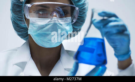 Aufmerksame biracial Chemiker in Erlenmeyerkolben mit blauer Flüssigkeit im Inneren suchen Stockfoto