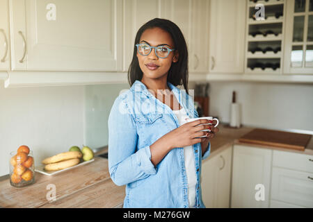 Lächelnde junge afrikanische Frau mit Brille und trinken eine Tasse frischen Kaffee im Stehen in Ihrer Küche Stockfoto