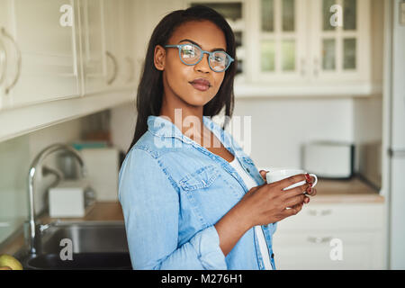 Selbstbewussten jungen afrikanischen Frau Brille trägt in ihrer Küche zu Hause trinken stehen eine Tasse frischen Kaffee Stockfoto