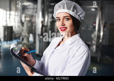 Ein hübsches junges Mädchen, Wissenschaftler oder Mitarbeiter in weiße Uniform macht sich Notizen auf Papier vor dem Hintergrund der modernen Fabrik. Stockfoto