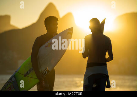 RIO DE JANEIRO - 20. März 2017: Sonnenuntergang Silhouetten von zwei junge Surfer mit Surfbrettern am Arpoador mit zwei Brüder Bergen im Hintergrund Stockfoto