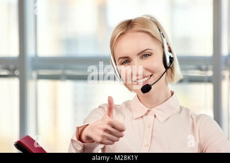 Weibliche blond reife Call Center Betreiber mit Daumen nach oben. Lächelnd Call center Agent Daumen hoch geben gegen Windows Hintergrund. Stockfoto