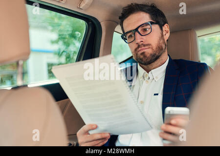 Junge Unternehmer lesen Schreibarbeiten und über ein Handy, während auf dem Rücksitz eines Autos durch die Stadt gefahren wird, sitzen Stockfoto