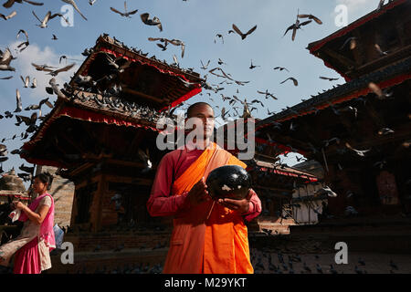 Nepalesische Mönch trägt orange robe Holding urn für Almosen am Durbar Square in Kathmandu Nepal, mit street Tauben fliegen Overhead, an einem sonnigen Tag. Stockfoto