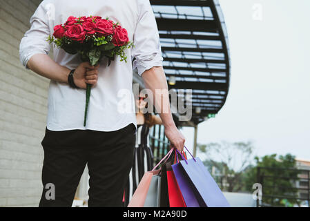 Mann, Shopping Bag & Strauß roter Rosen hinter seinem Rücken für Frau. Freund überraschen Freundin mit Blume im Valentinstag. ann Feiern Stockfoto