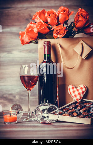 Rote Flasche Wein, eine Schachtel Pralinen, zwei Gläser Wein, Rosen in eine Papiertüte mit Deko von roten Herzen auf Holztisch. Valentines Tag Feier Stockfoto