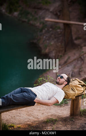 Mann draußen entspannen. Tragen ein weißes T-Hemd, dunkle Hose, Sonnenbrille, ist ein junger Mann auf Holzbank gegen See in einem Wald im freien liegend Stockfoto