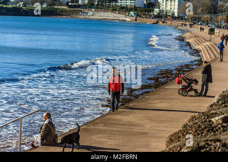 Menschen zu Fuß entlang der Promenade von Torre Strand bei Flut im Februar Sonnenschein. Nur 2 Grad C, aber viel der Leute aus trotzen der kalten aber sonnigen Morgen bei Torquay, Torbay, Devon, Großbritannien. Februar 2018 Stockfoto