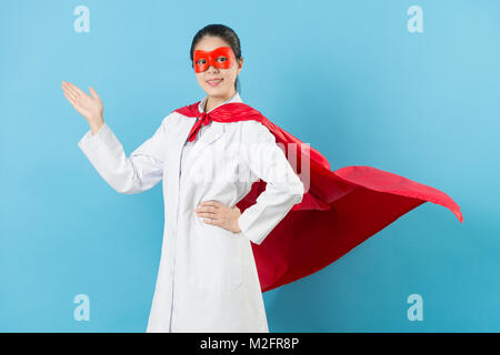 Vertrauen hübsche Frau Klinik Arzt tragen Superhelden Bekleidung Gesicht zur Kamera und Geste auf blauem Hintergrund vorstellen. Stockfoto