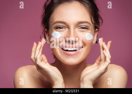 Lachende Mädchen Anwendung Feuchtigkeitscreme auf ihrem Gesicht. Foto der jungen Mädchen mit makelloser Haut auf rosa Hintergrund. Hautpflege und Schönheit Konzept Stockfoto