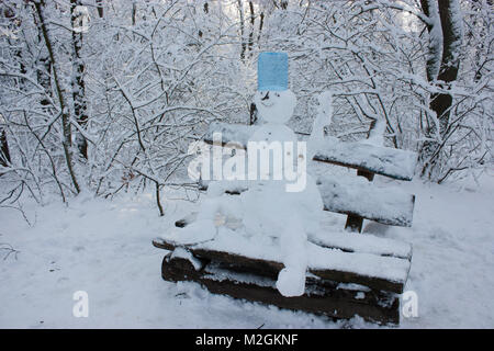 Schneemann sitzt auf einer Bank im Wald im Winter, viel Schnee, Bäume im Schnee Stockfoto