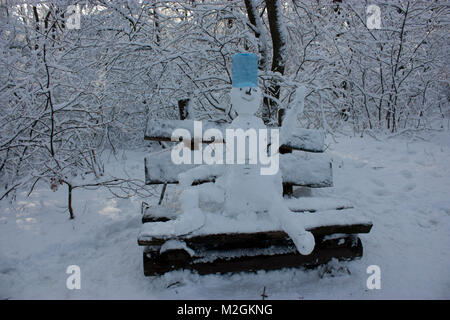 Schneemann sitzt auf einer Bank im Wald im Winter, viel Schnee, Bäume im Schnee Stockfoto