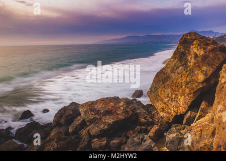 Sonnenuntergang entlang Point Dume State Beach mit bunten Himmel, Wellen, die in Felsformationen, und dem Santa Monica Berge in der Ferne, Ma Stockfoto