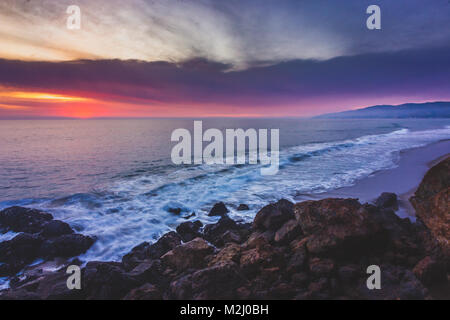 Sonnenuntergang entlang Point Dume State Beach mit bunten Himmel, Wellen, die in Felsformationen, und dem Santa Monica Berge in der Ferne, Ma Stockfoto