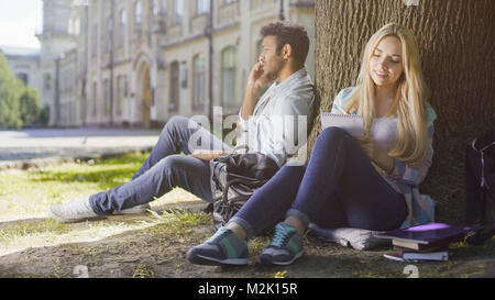 Multirassischen Kerl Gespräch am Handy unter Baum, Mädchen Notizen im Notebook Stockfoto