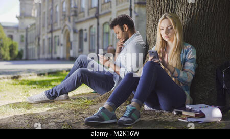 Weibliche, männliche Fremde sitzen unter Bäumen, mit Handy, Mädchen schauen besorgt Stockfoto