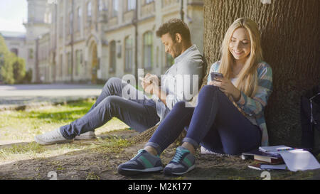 Junge Menschen sitzen unter Bäumen, mit Handy, auf sich selbst zu Lächeln, Freude, Filmmaterial Stockfoto